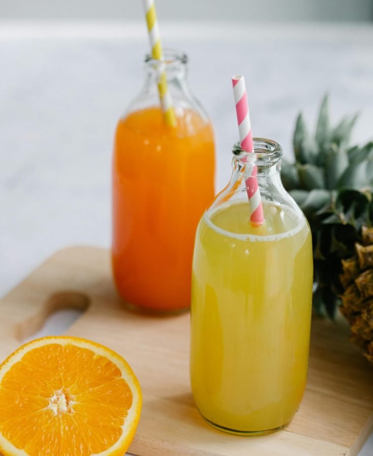 Two glass bottles of fresh citrus and pineapple juices with colorful straws on a wooden board.